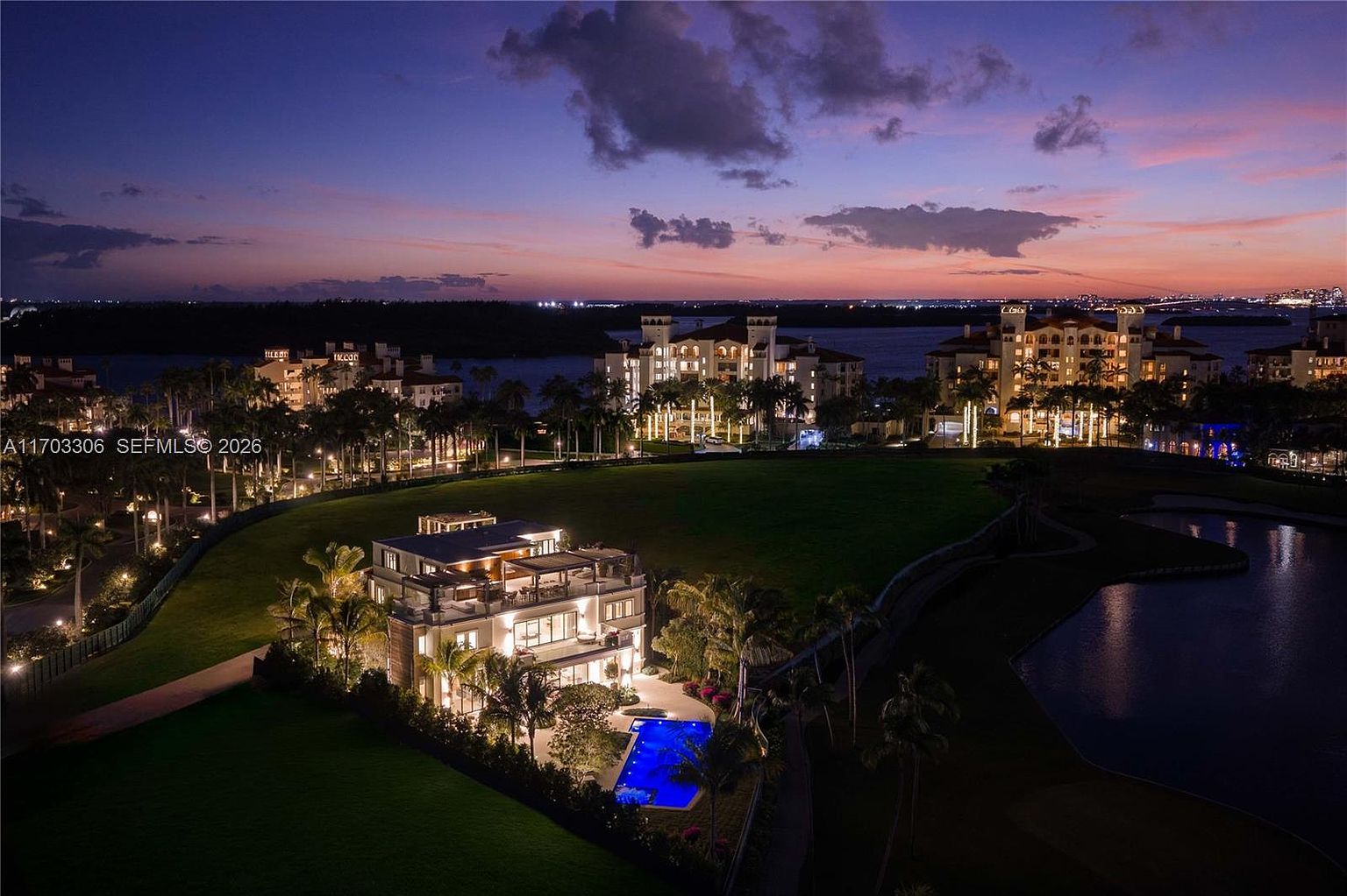 This stunning aerial twilight shot captures a luxurious multi-story estate set against a vibrant sunset sky. The property features a beautifully illuminated swimming pool, expansive manicured lawns, and a modern architectural design with multiple terraces. In the background, a serene body of water and distant city lights create a sophisticated and tranquil atmosphere.