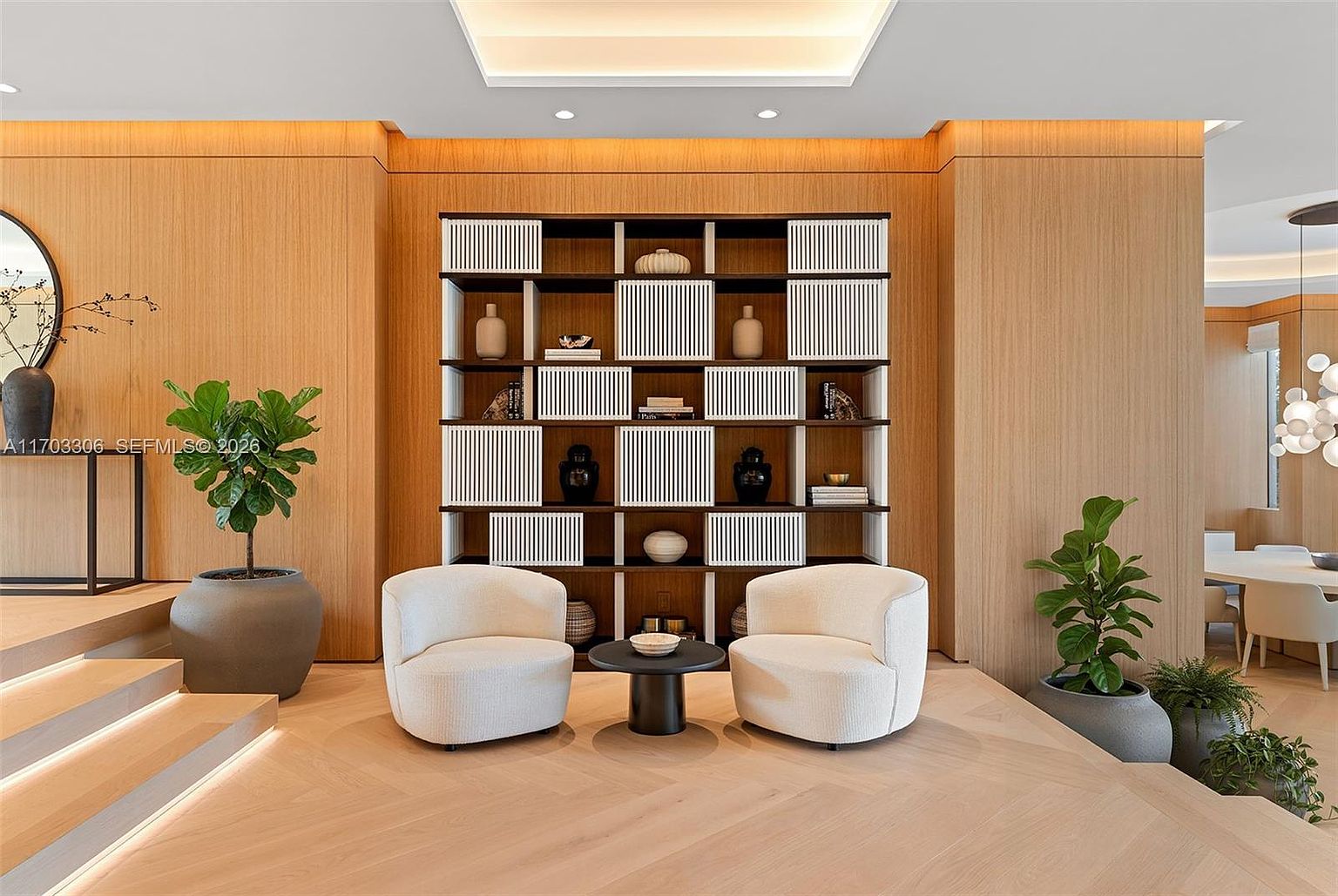 This sophisticated living area features a striking floor-to-ceiling wooden accent wall with a built-in shelving unit, creating a warm and modern focal point. Two plush, cream-colored curved armchairs are positioned around a small, dark circular side table, resting on light-toned herringbone wood flooring. The space is accented by lush potted plants and soft, recessed lighting, conveying an atmosphere of refined luxury and comfort.