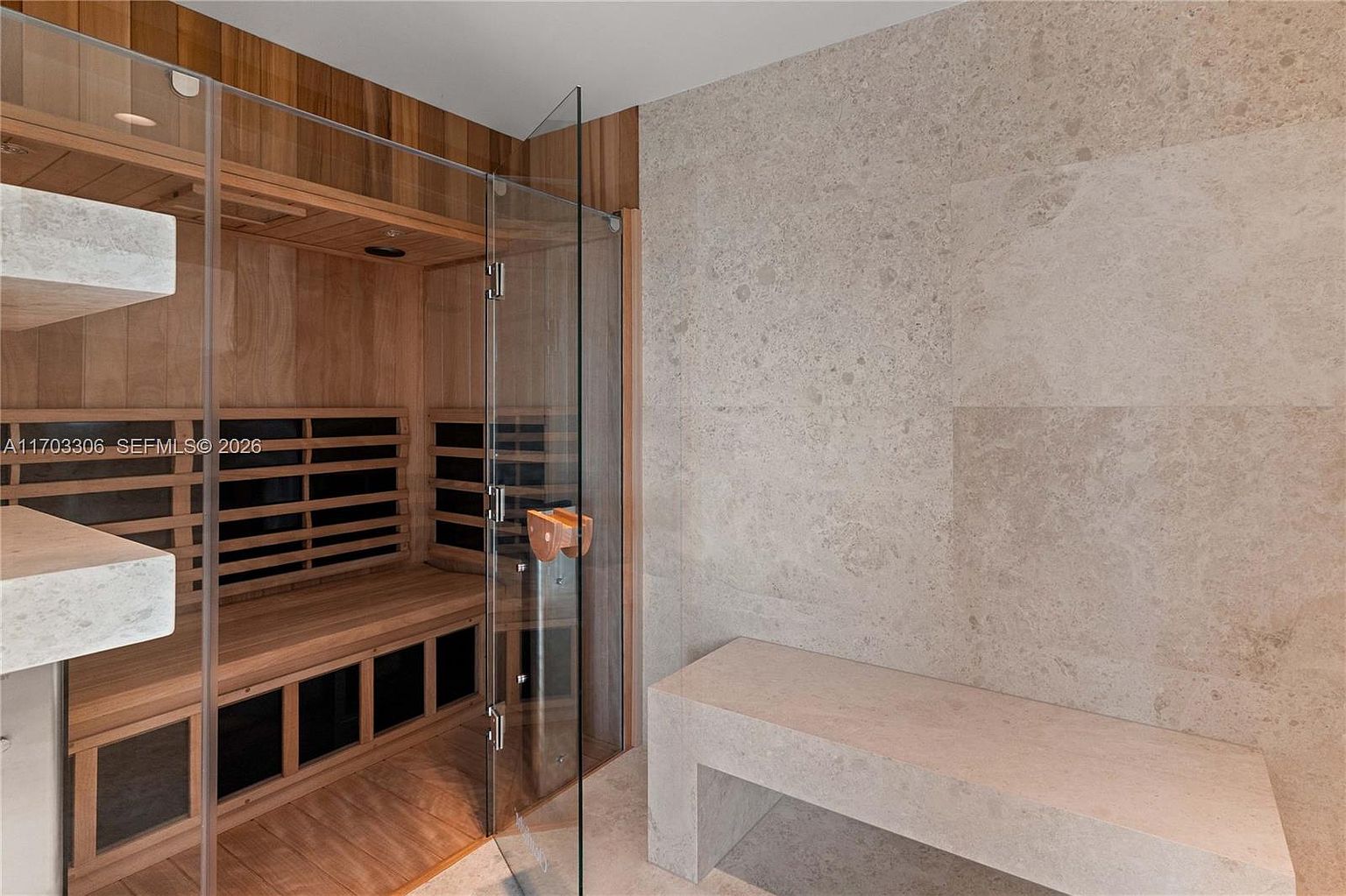 This luxurious spa-inspired bathroom features a custom-built cedar sauna enclosed in glass, positioned next to a minimalist stone bench. The room is finished with high-end, neutral-toned stone tiles that create a serene and sophisticated atmosphere. The perspective is eye-level, highlighting the seamless integration of wellness amenities within a modern residential space.
