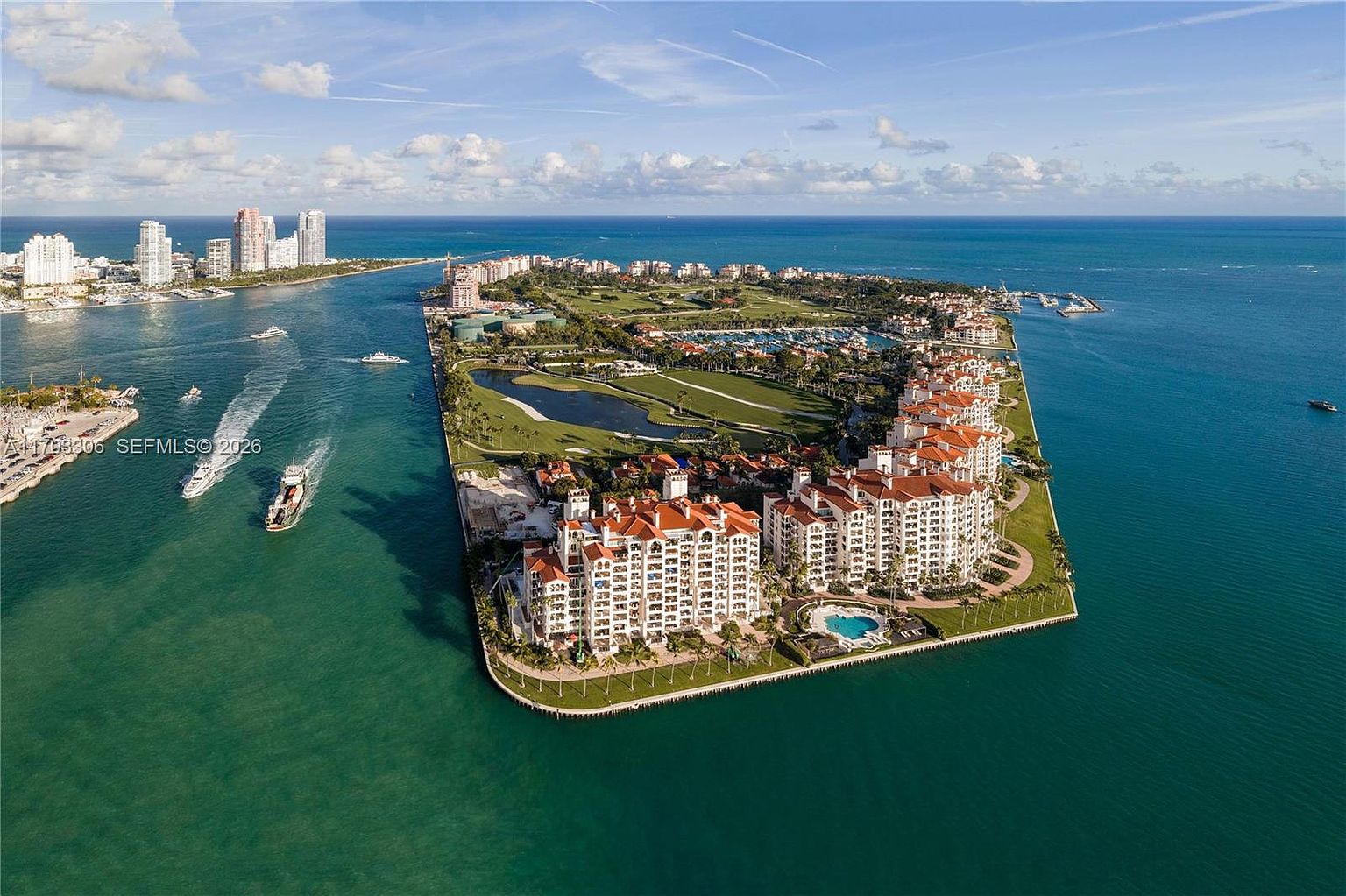 This stunning aerial view captures a luxury waterfront residential complex situated on a peninsula, surrounded by vibrant turquoise waters. The property features Mediterranean-style architecture with terracotta roofs, lush landscaping, and a private pool area, all set against the backdrop of a golf course and the open ocean. The perspective provides a comprehensive look at the prime location, highlighting the exclusive island-like lifestyle and proximity to the water.