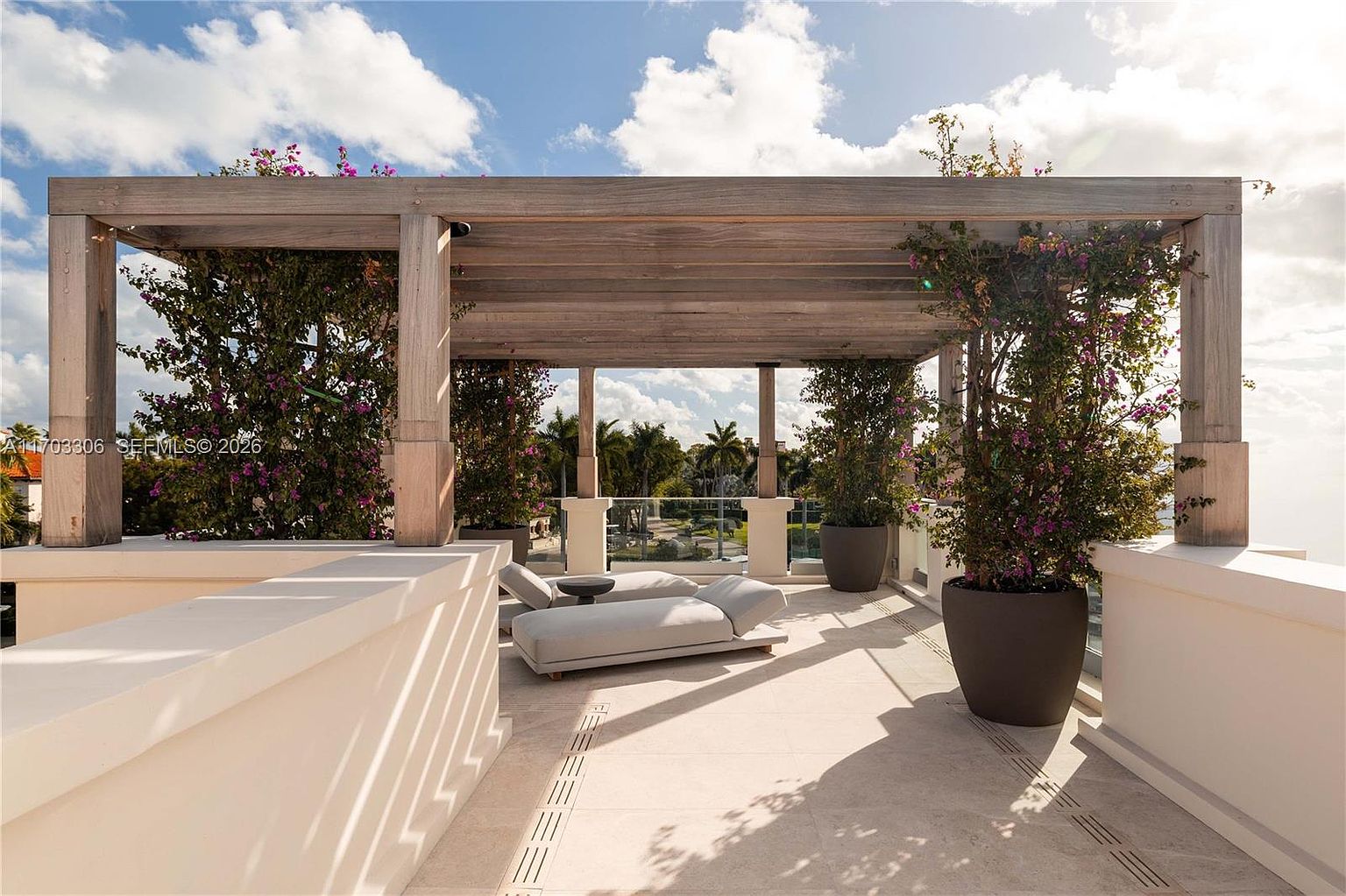 This luxurious rooftop terrace features a wooden pergola providing partial shade over a pair of modern lounge chairs. The space is adorned with lush potted bougainvillea plants, creating a vibrant and private outdoor retreat with a clear view of the surrounding landscape. The clean, light-toned flooring and minimalist design offer a sophisticated and serene atmosphere perfect for relaxation.