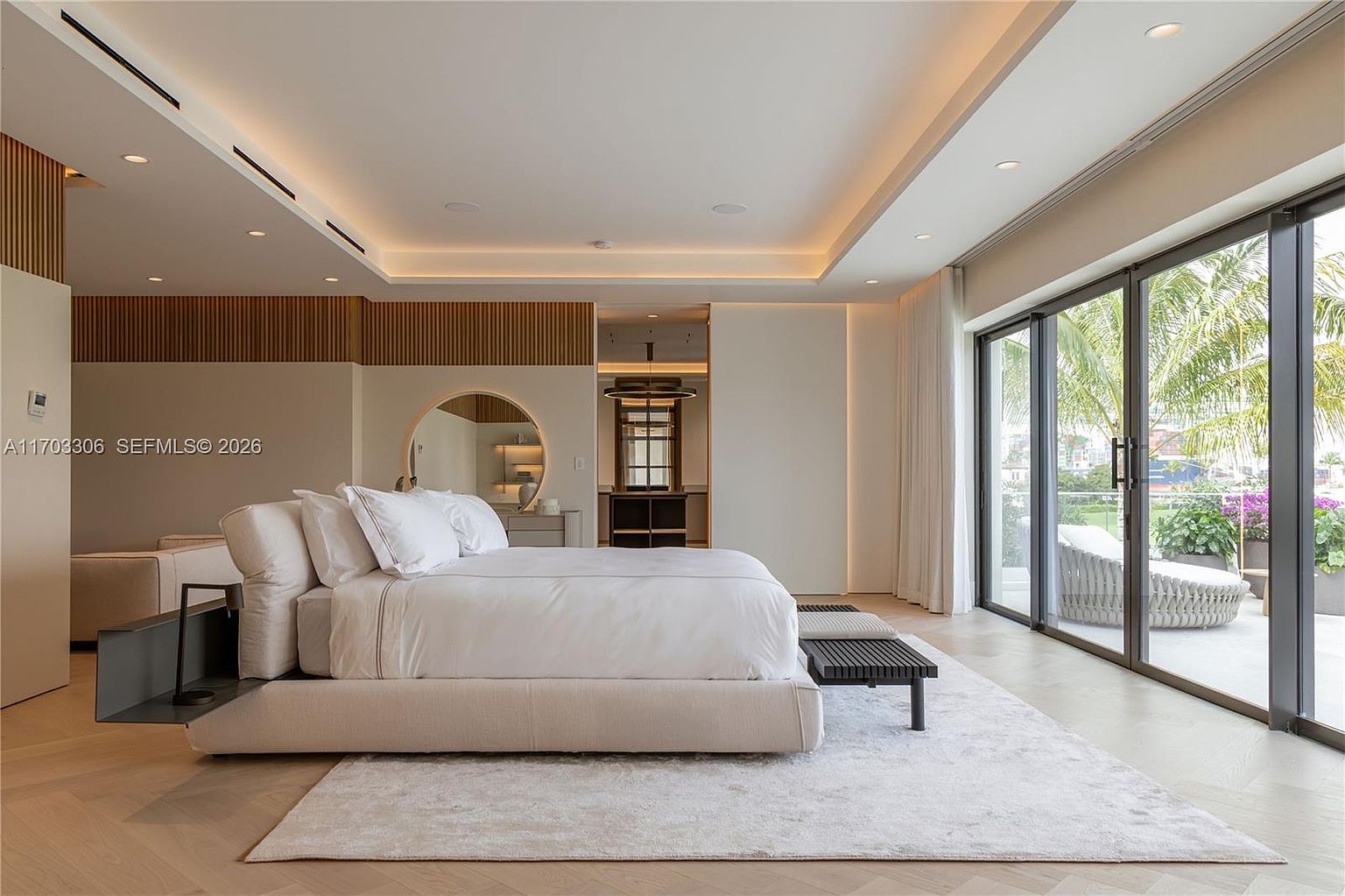 This luxurious primary bedroom features a minimalist, contemporary design with a neutral color palette and warm ambient cove lighting. A plush, low-profile bed serves as the centerpiece, complemented by a sleek wooden bench and a large circular mirror, while floor-to-ceiling glass sliding doors provide a seamless transition to a private outdoor terrace. The room exudes a serene, high-end atmosphere, enhanced by clean lines and sophisticated architectural detailing.