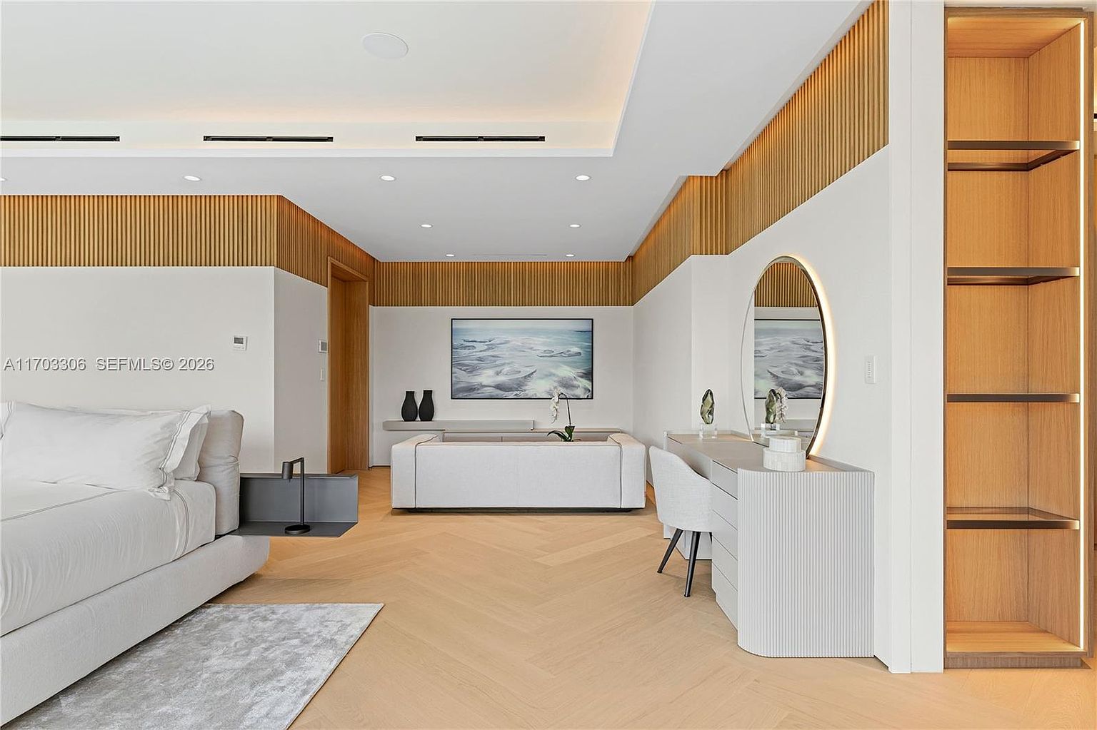This luxurious primary bedroom features a sophisticated, minimalist design with light wood herringbone flooring and warm wood-slat wall accents. The space is anchored by a plush, neutral-toned bed and a sleek vanity area with a backlit circular mirror, creating an atmosphere of refined elegance. A built-in shelving unit and a low-profile bench add functional style, while the soft, recessed lighting enhances the serene and high-end aesthetic.