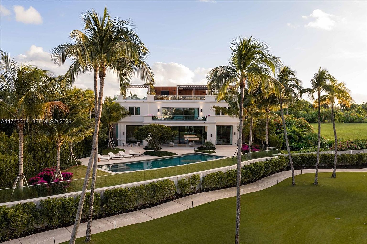 This stunning rear view of a luxury estate showcases a modern, multi-level architectural design with a pristine swimming pool and expansive patio area. The property is surrounded by lush landscaping, tall palm trees, and a manicured lawn, creating a serene and private atmosphere. The perspective is captured from an elevated angle, highlighting the seamless integration between the indoor living spaces and the outdoor recreational area.