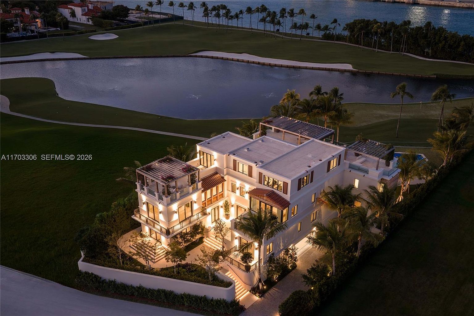This stunning aerial view captures a luxurious multi-story Mediterranean-style estate illuminated at dusk. The property features expansive terraces, a private rooftop deck, and lush landscaping, all set against the backdrop of a serene golf course and a tranquil lake. The warm lighting highlights the architectural elegance and sophisticated design of this high-end residence.