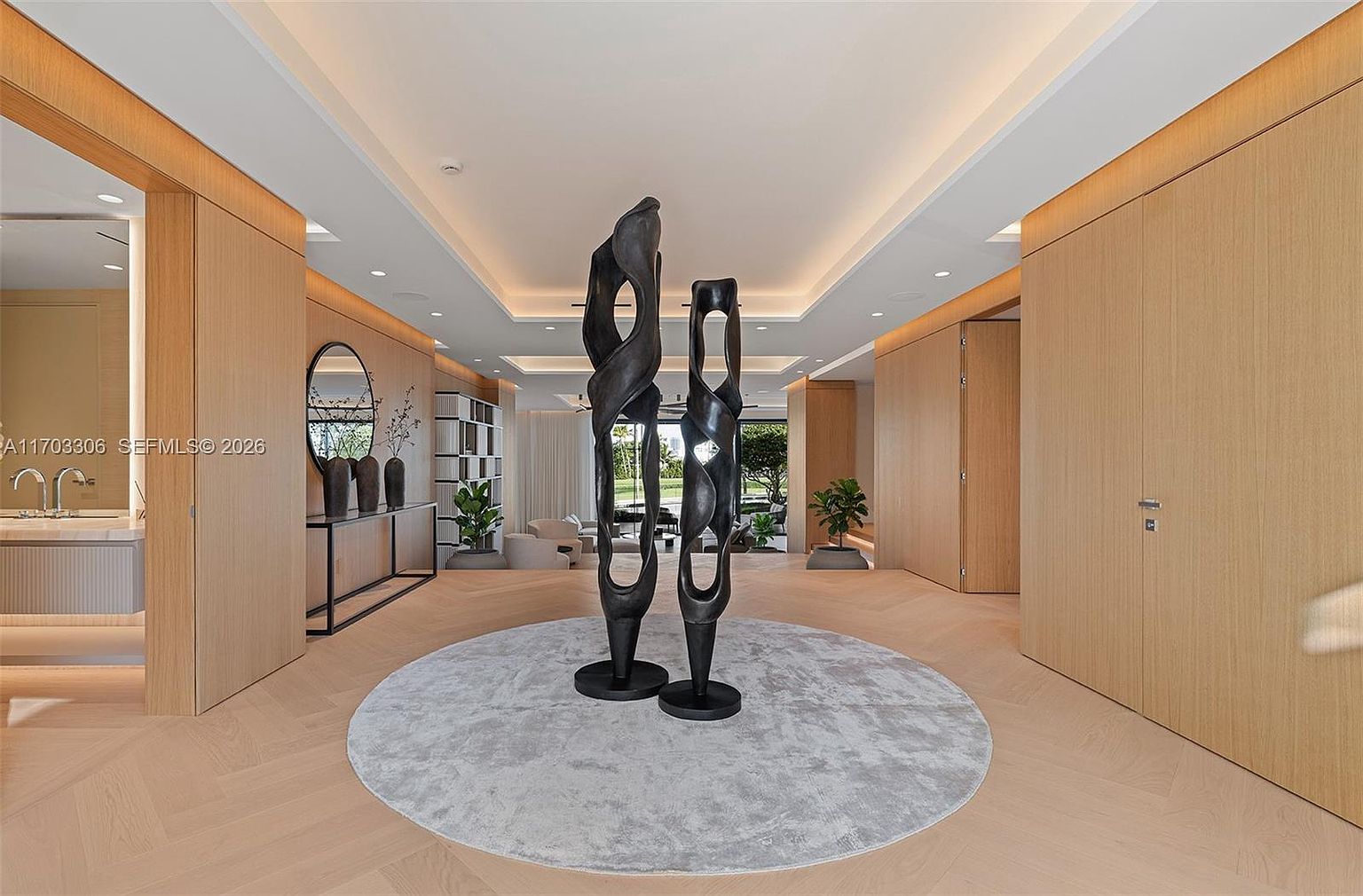 This sophisticated entryway features a grand, open-concept design with light wood flooring and warm, recessed cove lighting. A striking, tall black abstract sculpture serves as the focal point on a circular grey rug, leading the eye toward a bright, airy living space in the background. The clean lines and minimalist aesthetic create a luxurious, gallery-like atmosphere.