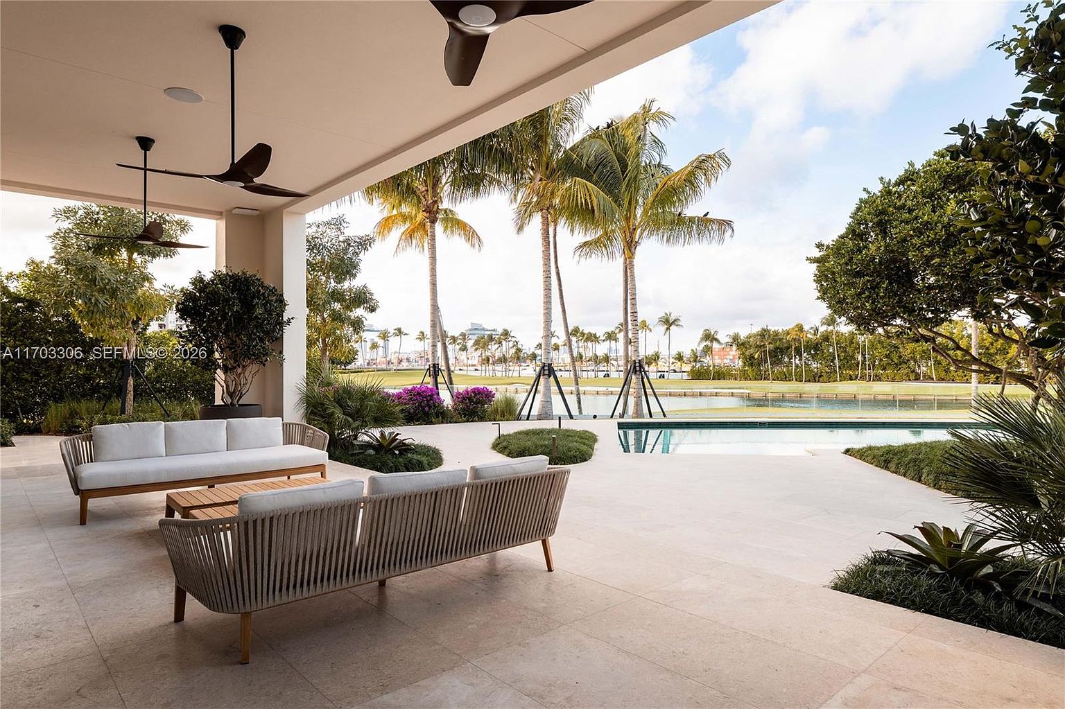 This luxurious covered patio offers a seamless transition to an outdoor oasis, featuring a modern sofa set and a sleek, minimalist design. The space overlooks a pristine swimming pool and a lush, manicured landscape with tall palm trees and a serene water view in the background. The open-air architecture and neutral stone flooring create an inviting, high-end atmosphere perfect for relaxation and entertaining.