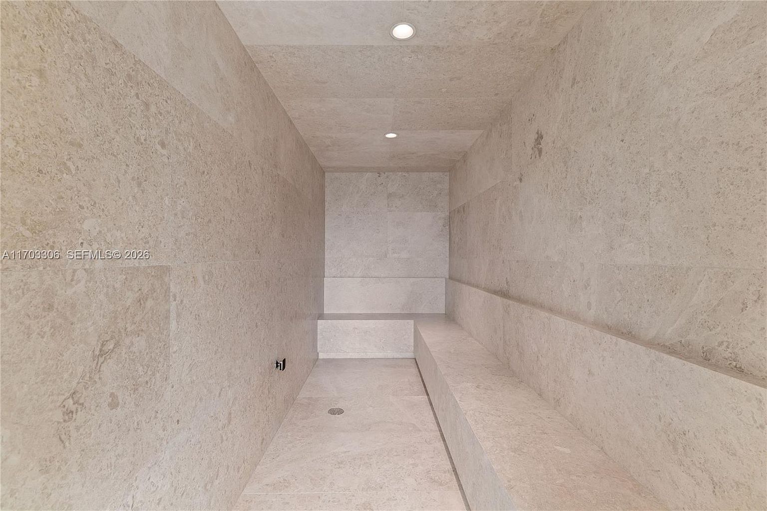 This image features a minimalist, stone-clad steam room or sauna, characterized by its monochromatic beige marble walls and built-in bench seating. The space is illuminated by recessed ceiling lights, creating a serene and spa-like atmosphere. The perspective is a straight-on, eye-level shot that emphasizes the clean lines and luxurious, uniform texture of the stone surfaces.