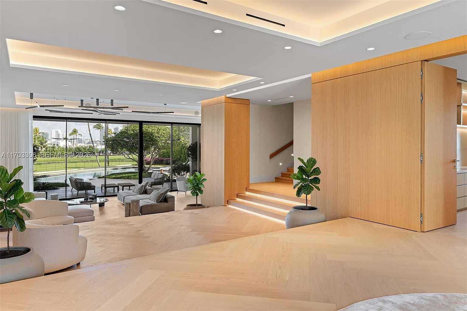 This expansive, modern living space features light-toned herringbone wood flooring and a seamless transition to an outdoor patio through floor-to-ceiling glass walls. The room is anchored by a neutral-toned seating area and accented with potted greenery, while a set of wooden stairs leads to an elevated level, creating a sophisticated, open-concept atmosphere. The design emphasizes clean lines, natural light, and a harmonious blend of indoor and outdoor living.