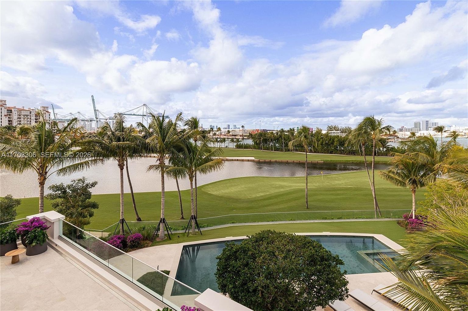 This elevated perspective captures a luxurious outdoor living space featuring a sleek, rectangular infinity-edge pool overlooking a pristine golf course and a tranquil lake. The scene is framed by lush palm trees and vibrant purple bougainvillea, with a distant cityscape and industrial cranes visible on the horizon under a dramatic, cloud-filled sky. The composition emphasizes a high-end, resort-style atmosphere, blending private relaxation with expansive, scenic views.