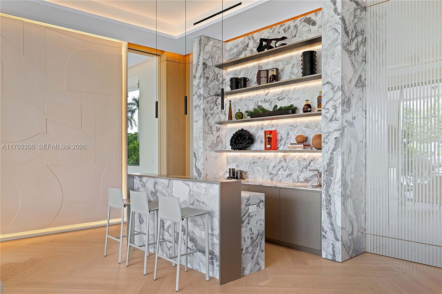 This sophisticated home bar area features striking marble countertops and backsplash, complemented by sleek, minimalist bar stools. The space is accented by open shelving displaying curated decor and warm, recessed lighting that creates an inviting, upscale atmosphere. The design seamlessly blends modern luxury with clean lines, making it a perfect focal point for entertaining.