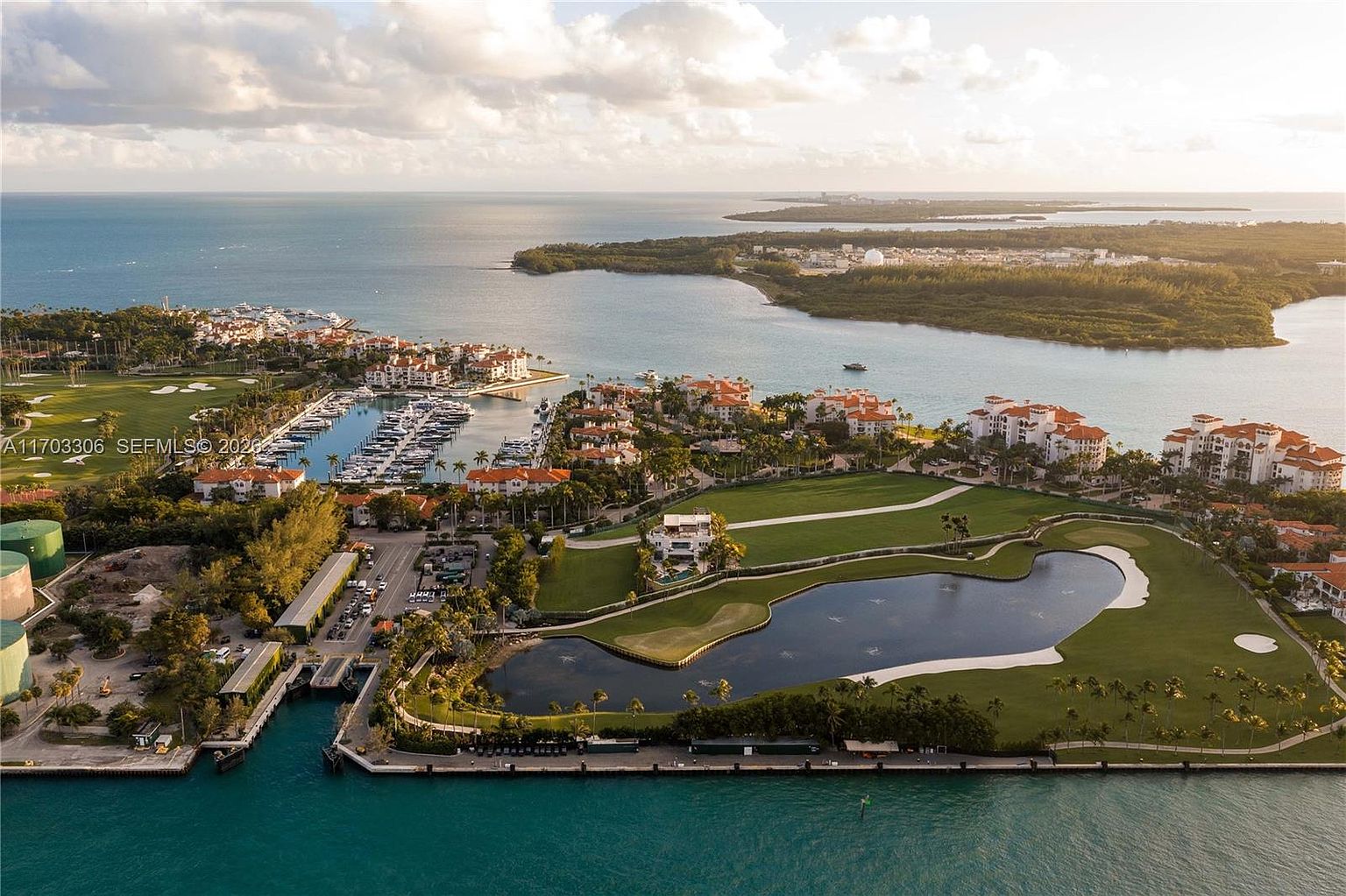 This stunning aerial view captures a luxurious waterfront residential community featuring a private marina, expansive green lawns, and a scenic golf course pond. The Mediterranean-style architecture with terracotta roofs blends seamlessly into the lush tropical landscape, all set against the backdrop of a vast, sparkling bay. The perspective provides a comprehensive look at the upscale coastal lifestyle and the prime location of the property.