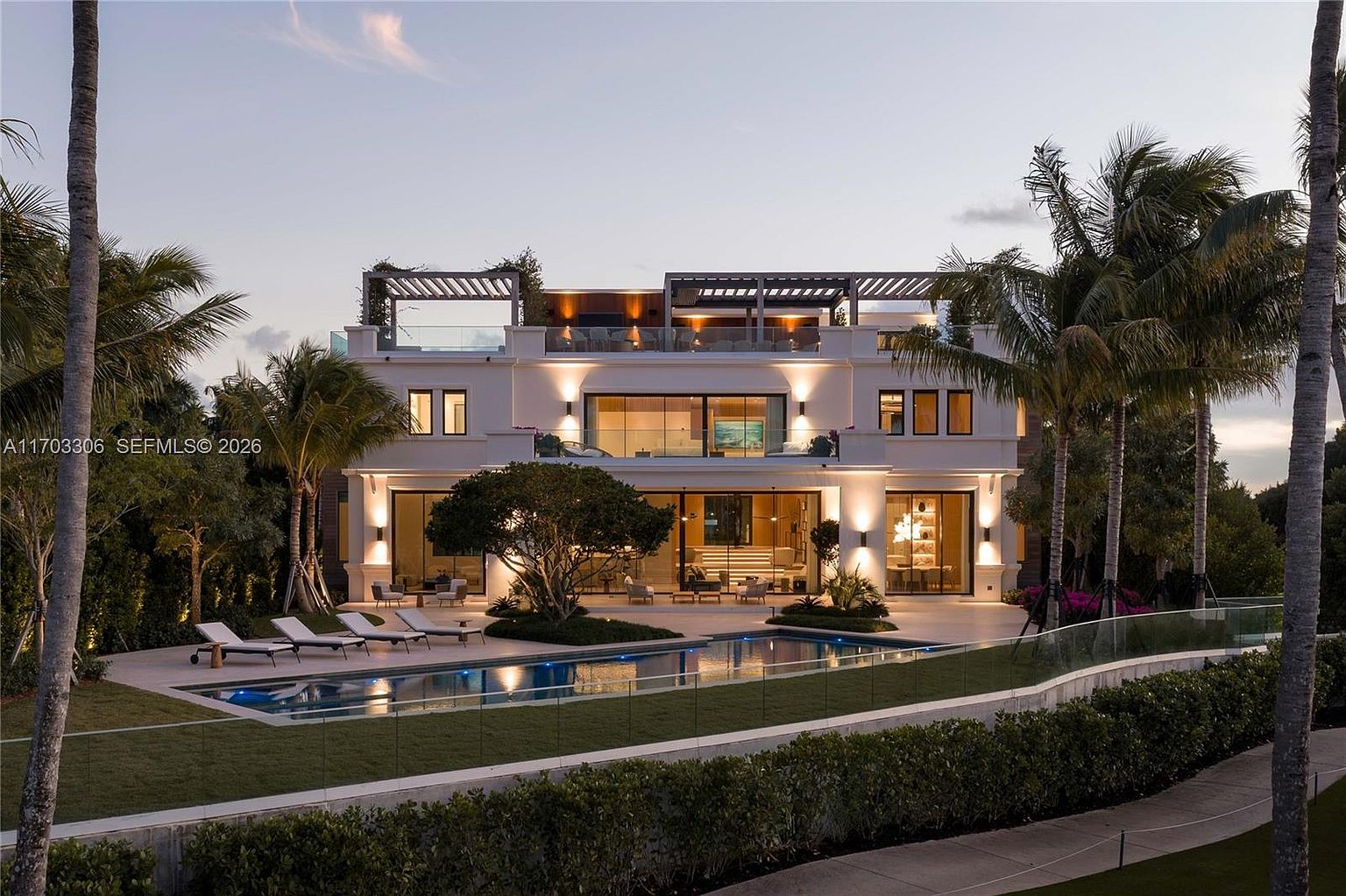 This stunning contemporary luxury estate is captured from a rear perspective at dusk, showcasing a multi-level architectural design with expansive glass walls and warm ambient lighting. The property features a pristine swimming pool surrounded by a manicured lawn, lounge chairs, and lush tropical landscaping, creating a sophisticated resort-like atmosphere. The cinematic composition highlights the seamless transition between the indoor living spaces and the outdoor entertainment area.