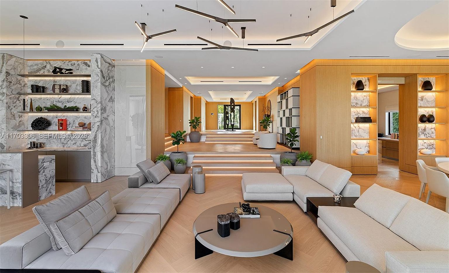 This expansive, ultra-modern living room features a sophisticated open-concept design with light wood flooring, a large modular sectional sofa, and a central circular coffee table. The space is highlighted by a multi-level layout with illuminated steps leading to a grand entryway, complemented by high-end marble shelving and unique geometric ceiling light fixtures. The overall aesthetic is one of luxurious, contemporary elegance with warm wood paneling and clean, minimalist lines.