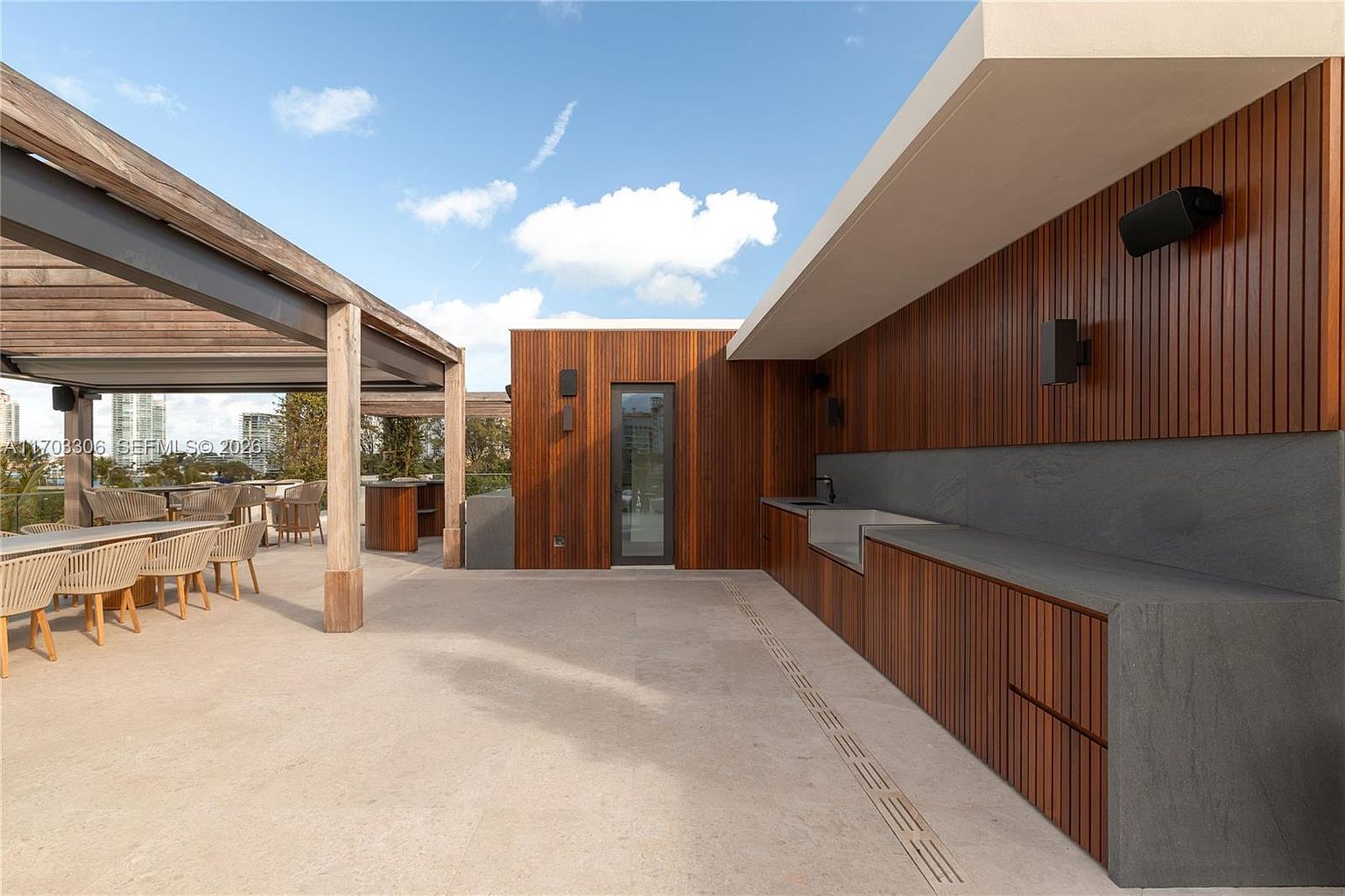 This sophisticated outdoor terrace features a sleek, modern design with warm wood-paneled walls and a minimalist stone countertop kitchen area. A pergola-covered dining space sits to the left, while the open-air layout provides a seamless transition for entertaining. The perspective is a wide-angle shot from the center of the deck, emphasizing the clean lines and high-end materials of this luxury rooftop space.