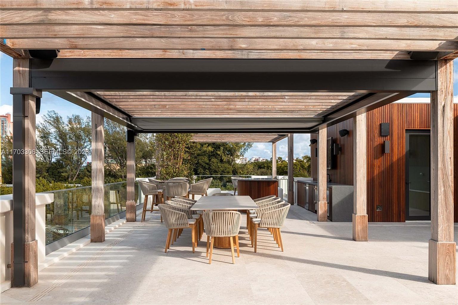 This expansive, modern rooftop terrace features a sophisticated pergola structure providing shade over a contemporary dining area with woven chairs and a sleek table. The space is complemented by a built-in outdoor kitchen with wood-paneled walls and glass railings that offer an unobstructed view of the surrounding lush greenery and distant skyline. The overall aesthetic is one of luxury, comfort, and seamless indoor-outdoor living, perfect for high-end entertaining.