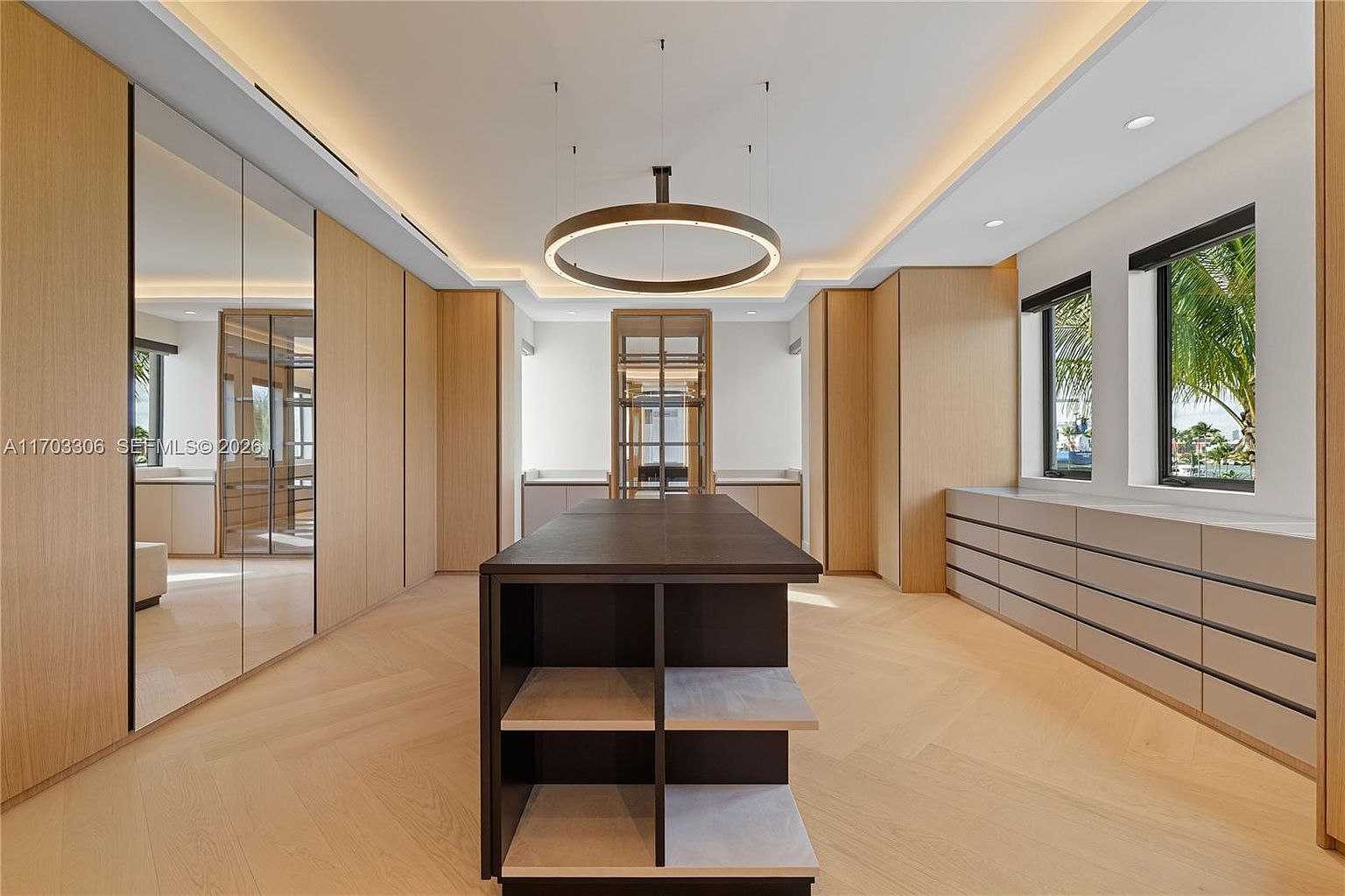 This luxurious, expansive walk-in closet features custom light wood cabinetry, a dark central island with open shelving, and a sophisticated circular pendant light. The room is finished with light-toned herringbone wood flooring and large mirrored wardrobe doors that enhance the sense of space. Two windows provide natural light, creating an airy and high-end atmosphere perfect for a primary suite.