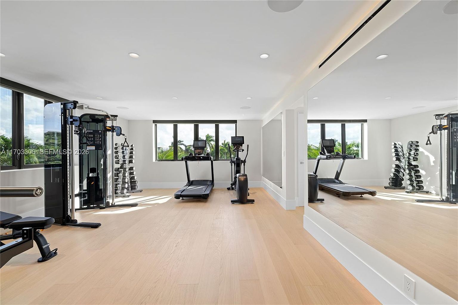 This spacious and bright home gym features light-toned hardwood flooring and a large wall-to-wall mirror that enhances the sense of space. The room is well-equipped with professional-grade fitness equipment, including a treadmill, an elliptical machine, and a multi-station strength training system, all positioned to overlook the outdoor greenery through large windows. The clean, minimalist aesthetic and neutral color palette create an inviting and motivating environment for a workout.