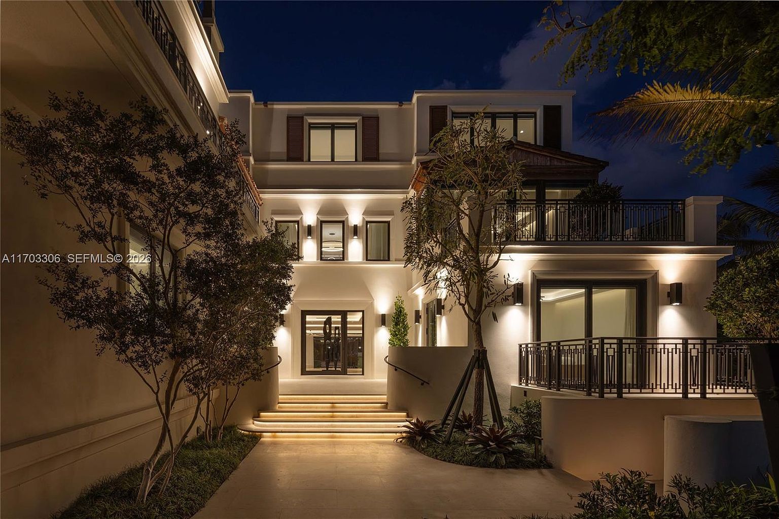 This stunning, modern luxury residence is captured at twilight, showcasing a grand, illuminated entryway with tiered, glowing steps leading to a sophisticated front door. The architectural design features clean lines, warm accent lighting, and a balanced composition of white stucco walls and dark metal railings. Lush landscaping and mature trees frame the structure, creating an inviting and high-end atmosphere perfect for a premium real estate listing.