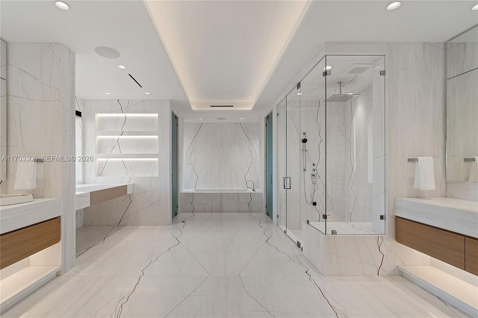 This luxurious primary bathroom features a symmetrical layout with dual floating vanities, a central soaking tub, and a spacious glass-enclosed walk-in shower. The room is finished in high-end white marble with dramatic veining, complemented by warm under-cabinet lighting and a recessed ceiling design. The overall aesthetic is ultra-modern, clean, and sophisticated, creating a spa-like atmosphere.