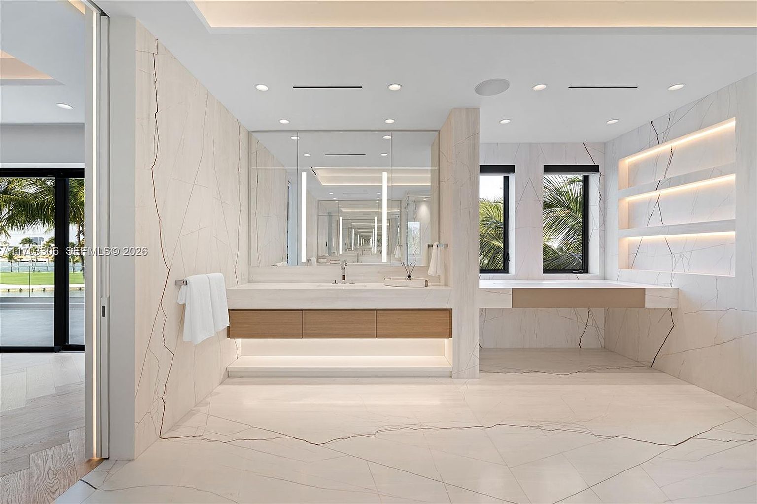 This luxurious primary bathroom features a sleek, modern design with floor-to-ceiling marble walls and matching flooring. The space includes a floating double vanity with wood-toned cabinetry, integrated lighting, and a large mirror, complemented by built-in recessed shelving on the right. A large window provides natural light and a view of the outdoors, creating an airy and sophisticated atmosphere.