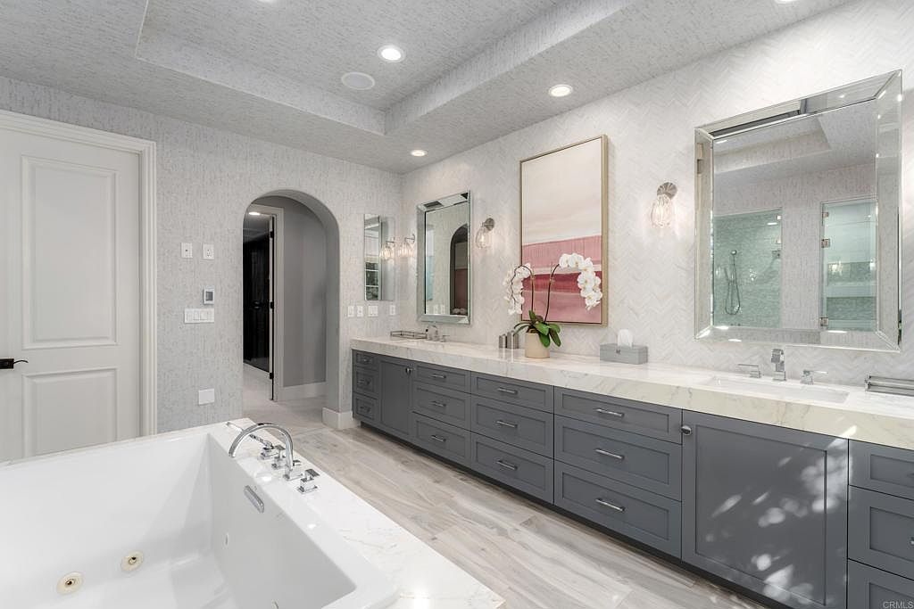 This is a luxurious primary bathroom featuring a large soaking tub in the foreground and a long vanity with gray cabinetry and a white countertop. The walls are covered in a textured wallpaper, and there are two large mirrors above the vanity, reflecting the spaciousness of the room. The overall impression is one of elegance and tranquility.