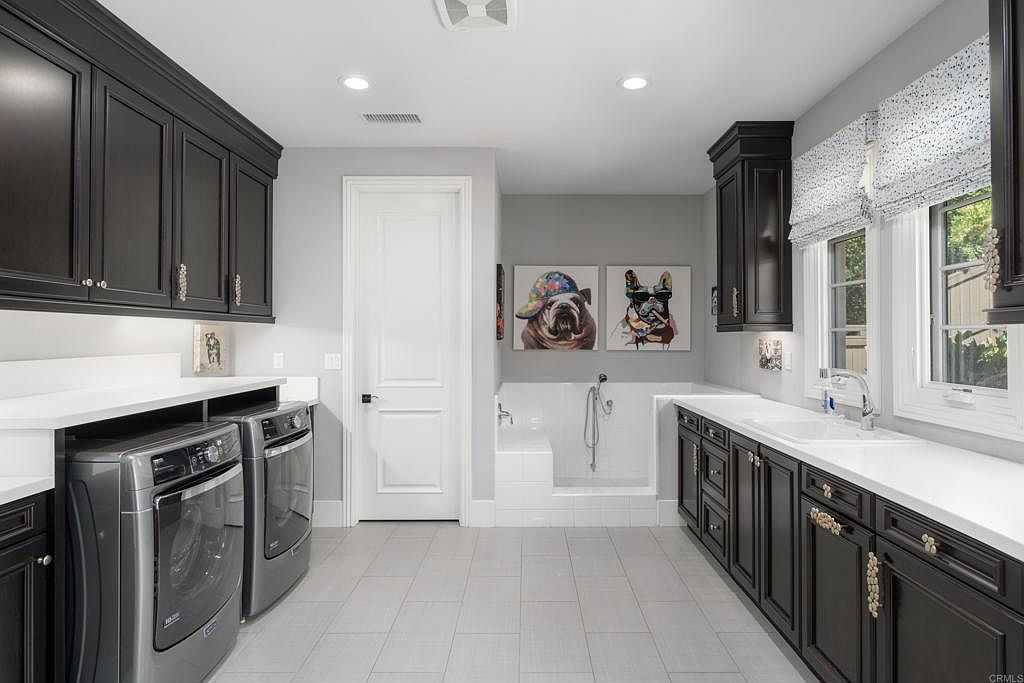 This is a well-appointed laundry room featuring dark cabinetry with decorative hardware, white countertops, and modern gray front-loading washer and dryer. A unique feature is the built-in dog washing station, adding a touch of luxury and practicality. The room is brightly lit with recessed lighting and natural light from a window with a patterned roman shade.