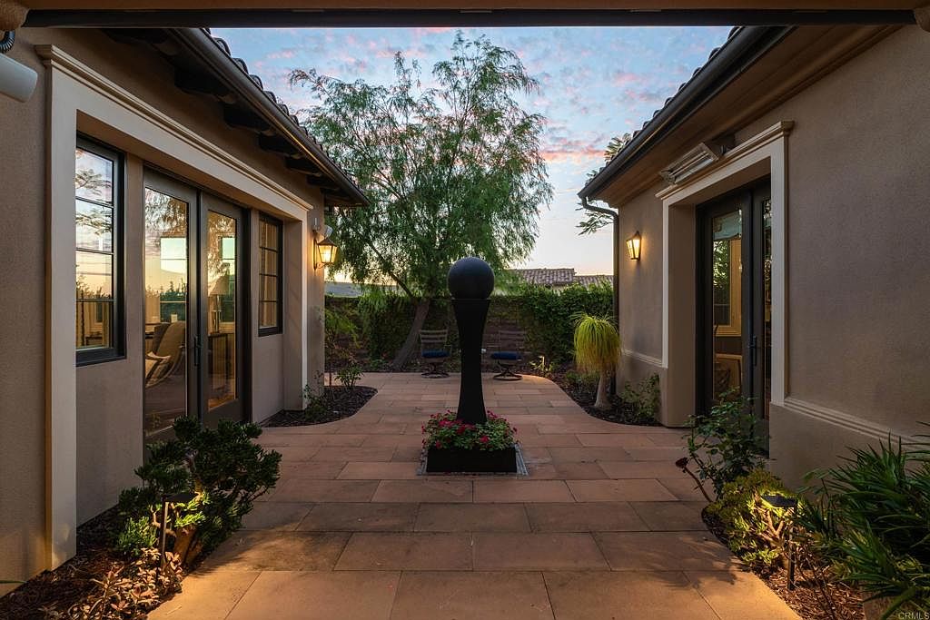 This image showcases a beautifully landscaped courtyard or garden area between two buildings. The space features a paved walkway, lush greenery, and a decorative water feature or sculpture in the center. The architecture is elegant with warm-toned exteriors and well-lit entryways, creating an inviting and serene atmosphere.