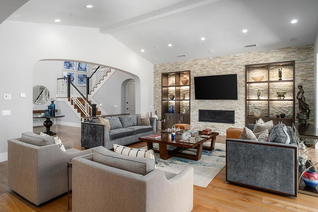 This is an interior shot of a living room featuring hardwood floors, a stone accent wall with a fireplace and built-in shelving units, and comfortable seating arrangements including sofas and armchairs. The room is well-lit with recessed lighting and natural light, creating a warm and inviting atmosphere. The camera is positioned to capture the entire living space, showcasing the layout and design elements.