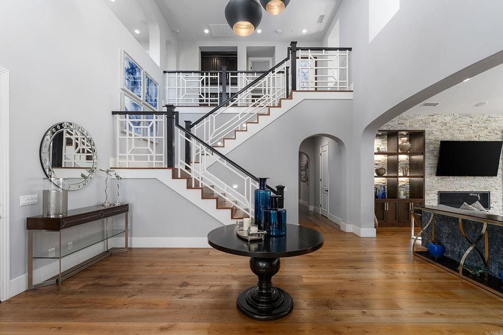 This interior shot showcases a grand foyer with a striking staircase featuring dark wood and white railings. A round table with blue vases sits in the center, adding a pop of color. The hardwood floors and neutral wall colors create a sophisticated and inviting atmosphere, while the architectural details and lighting fixtures enhance the luxurious feel of the home.