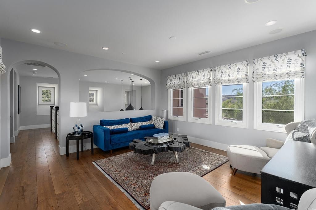 This is an interior shot of a living room featuring hardwood floors, a blue sofa, and a unique coffee table. The room is well-lit with natural light coming through the windows, which are adorned with patterned blinds. The space is decorated in a modern style with neutral wall colors and an area rug that adds warmth to the room.