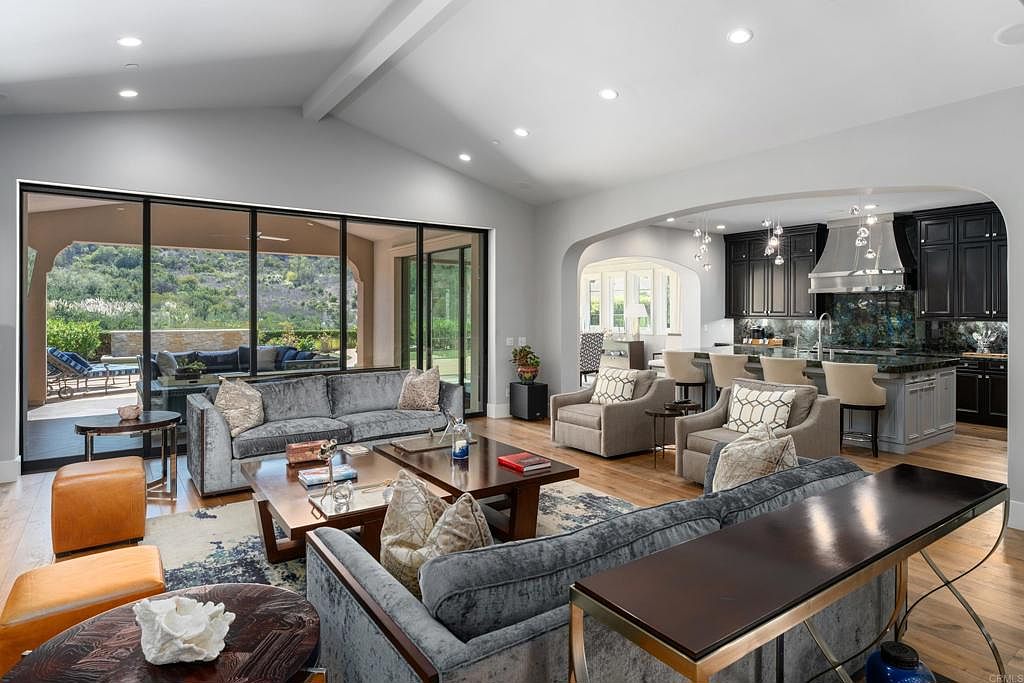 This is an interior shot of a luxurious living room that seamlessly transitions into a modern kitchen. The living room features plush grey sofas, wooden coffee tables, and large sliding glass doors that offer a view of the outdoor patio and landscape. The kitchen boasts dark cabinetry, a stainless steel range hood, and bar seating, creating an open and inviting space perfect for entertaining.