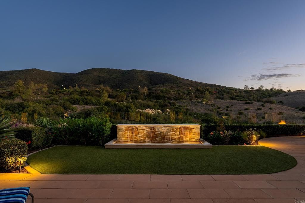 This image showcases a beautifully landscaped backyard featuring a modern water fountain as a focal point. The manicured lawn and surrounding greenery create a serene and inviting atmosphere. The patio area suggests ample space for outdoor entertaining, with a scenic mountain backdrop enhancing the property's appeal.