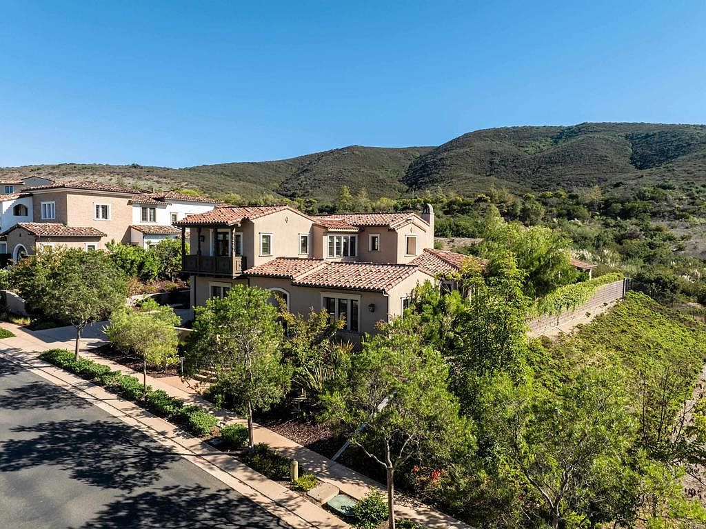 This aerial view showcases a beautiful two-story home with a red tile roof, set against a backdrop of rolling hills and clear blue skies. Lush greenery surrounds the property, enhancing its curb appeal and creating a sense of privacy. The house is part of a well-maintained neighborhood, suggesting a desirable location.