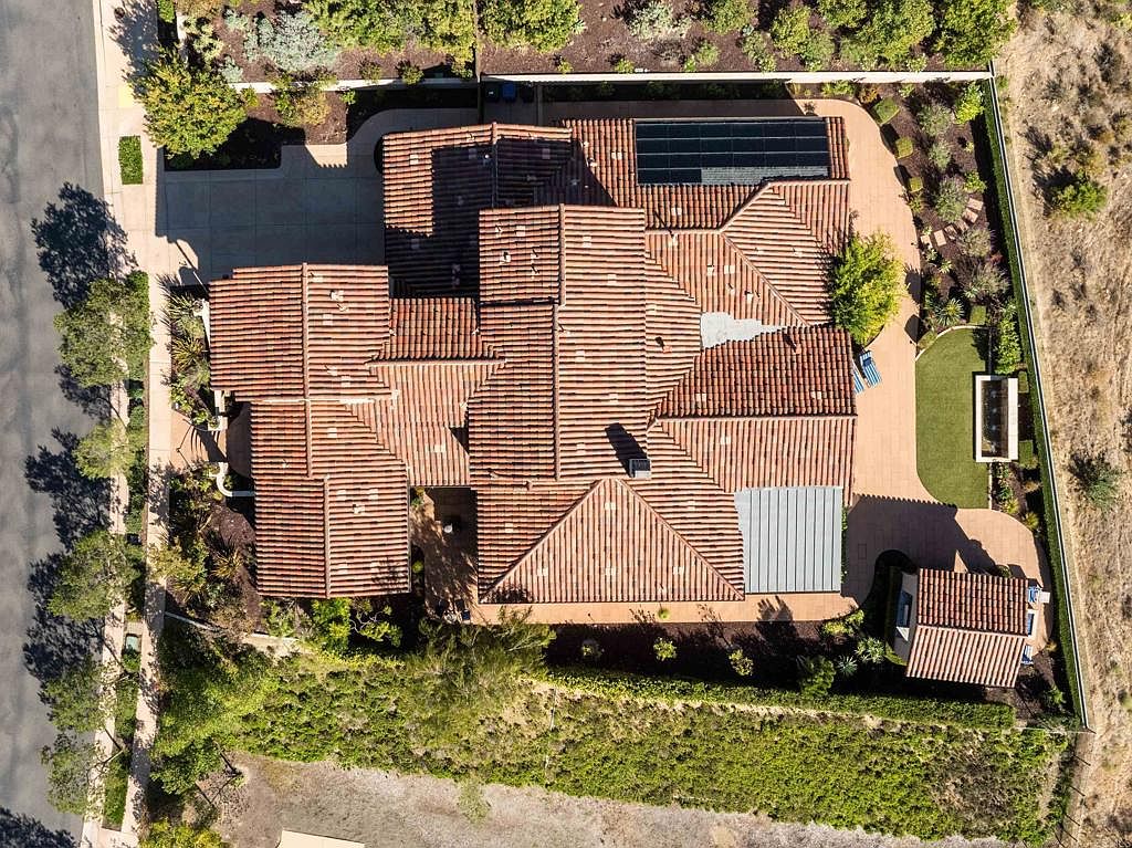 This aerial shot showcases a sprawling residential property with a terracotta tile roof, solar panels, and meticulously landscaped grounds. The house features multiple wings and a combination of traditional and modern architectural elements. A rectangular pool and paved walkways add to the property's appeal, creating a luxurious and private outdoor space.
