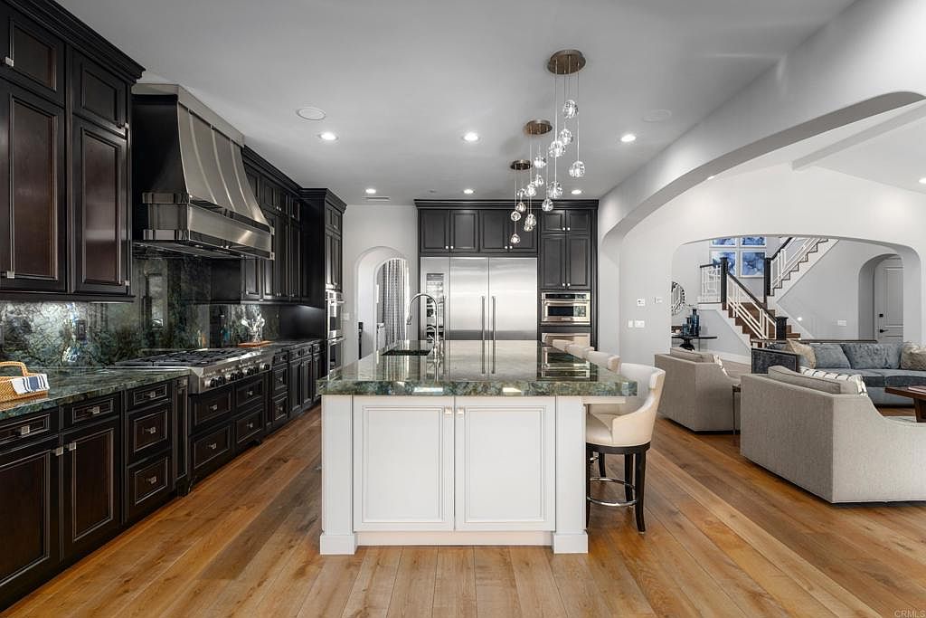 This is a luxurious kitchen featuring dark wood cabinetry, stainless steel appliances, and a large island with a light-colored countertop. The kitchen opens into a living area with hardwood floors and comfortable seating. The overall impression is one of elegance and spaciousness.