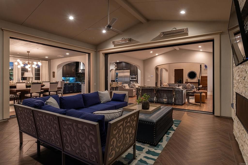 This is an interior shot of a luxurious living room that seamlessly connects to other areas of the home through wide openings. The room features a stylish blue sofa with decorative wooden backing, a plush ottoman, and a patterned area rug over hardwood flooring. The open layout allows for views into the dining room and another living space, creating a sense of spaciousness and connectivity.