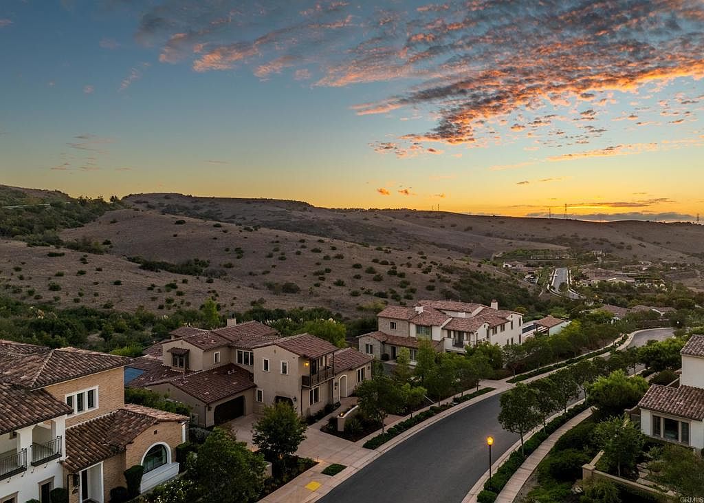 This aerial view showcases a collection of upscale homes with terracotta tile roofs nestled in a hillside community. The homes are surrounded by lush landscaping and a winding road, with a backdrop of rolling hills and a vibrant sunset sky. The image conveys a sense of luxury, tranquility, and desirable location.