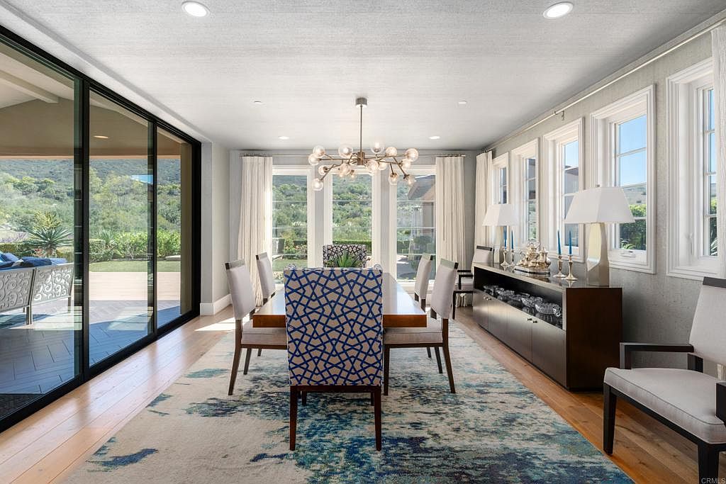 This is a bright and airy dining room featuring a long wooden table with upholstered chairs, one with a geometric pattern. A modern chandelier hangs above the table, and large windows and sliding glass doors offer views of the outdoor landscape. The room is decorated with a blue and white patterned rug and a dark wood sideboard, creating a sophisticated and inviting atmosphere.
