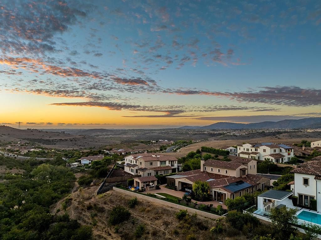 This aerial view showcases a collection of upscale homes nestled in a hillside community. The properties feature terracotta roofs and stucco exteriors, complemented by lush landscaping and private pools. The scene is set against a backdrop of rolling hills and a vibrant sunset sky, creating a sense of luxury and tranquility.