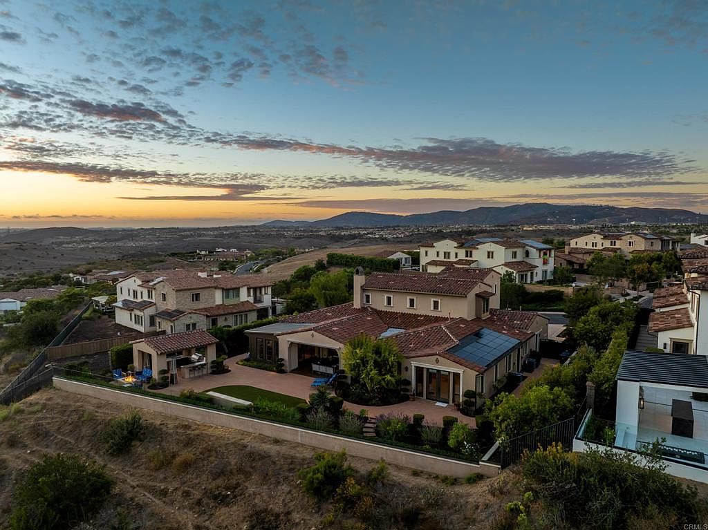 This aerial shot showcases a luxurious residential neighborhood at sunset, featuring multiple houses with red tile roofs and well-manicured landscaping. The houses are nestled on a hillside, offering scenic views of the distant mountains and the surrounding landscape. Solar panels are visible on one of the houses, adding a modern touch.