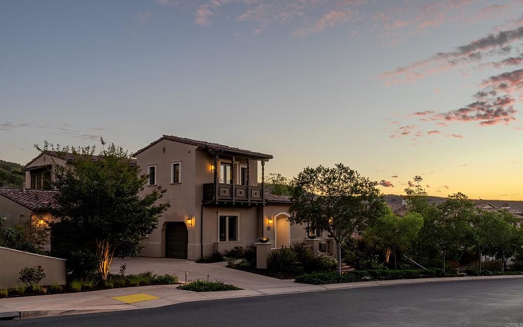 This is a front exterior view of a two-story Mediterranean-style home at dusk. The house features a stucco facade, a tile roof, and a small balcony. Landscaping includes trees and shrubs, and the overall impression is one of upscale suburban living. The perspective is from street level, capturing the home's curb appeal.