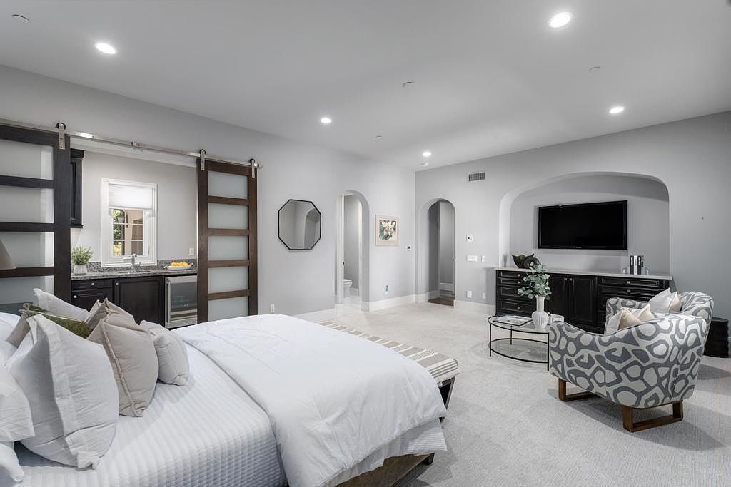 This is a spacious primary bedroom featuring a large bed with white linens and multiple pillows. The room includes a wet bar area with dark cabinetry and a wine fridge, as well as a seating area with a patterned armchair and a built-in entertainment center. The color palette is neutral, creating a serene and luxurious atmosphere.