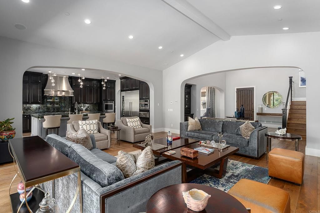 This is an interior shot of a living room that seamlessly transitions into a kitchen area. The living room features two plush sofas, a patterned area rug, and dark wood coffee tables, while the kitchen boasts dark cabinetry, stainless steel appliances, and bar seating. The open-concept design creates a spacious and inviting atmosphere, perfect for modern living.