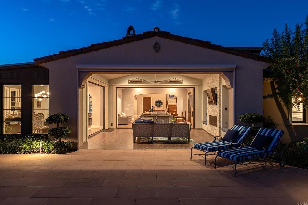 This image showcases a luxurious outdoor patio area extending from a home, featuring a seamless transition between indoor and outdoor living. The patio is furnished with comfortable lounge chairs and a stylish outdoor sofa, inviting relaxation. The open layout provides a clear view into the home's interior, highlighting the spaciousness and design continuity, while the warm lighting creates an inviting ambiance.