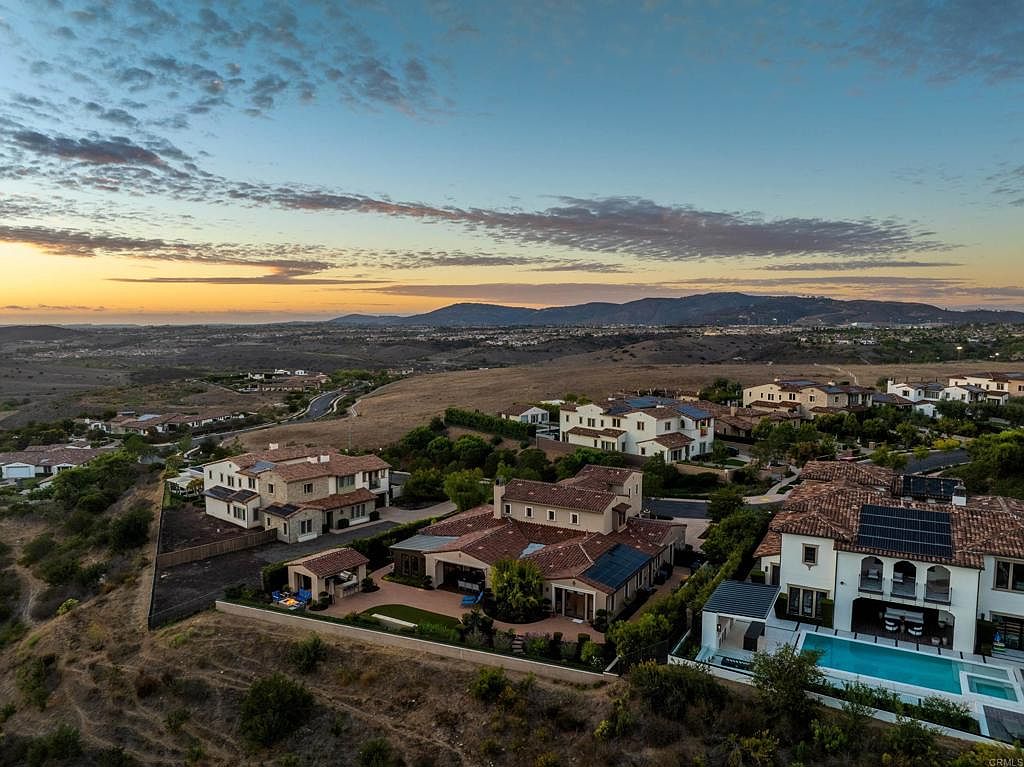 This aerial view showcases a luxurious residential neighborhood at sunset, featuring large, well-maintained homes with red tile roofs and private pools. The landscape includes rolling hills and distant mountains, creating a serene and upscale atmosphere. Solar panels are visible on some roofs, highlighting energy efficiency.