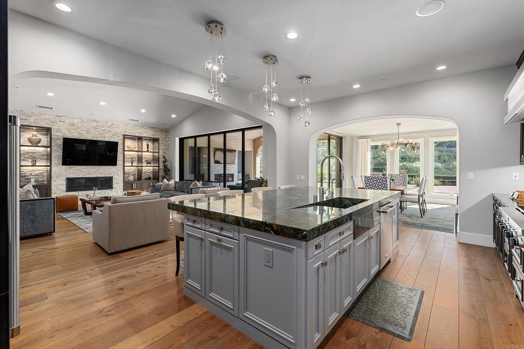 This interior shot showcases a modern, open-concept kitchen that seamlessly blends into the living and dining areas. The kitchen features a large island with a unique countertop, gray cabinetry, and pendant lighting, while the living room boasts a stone accent wall, built-in shelving, and comfortable seating. The dining area is bright and airy, with large windows offering views of the outdoors, creating a cohesive and inviting living space.