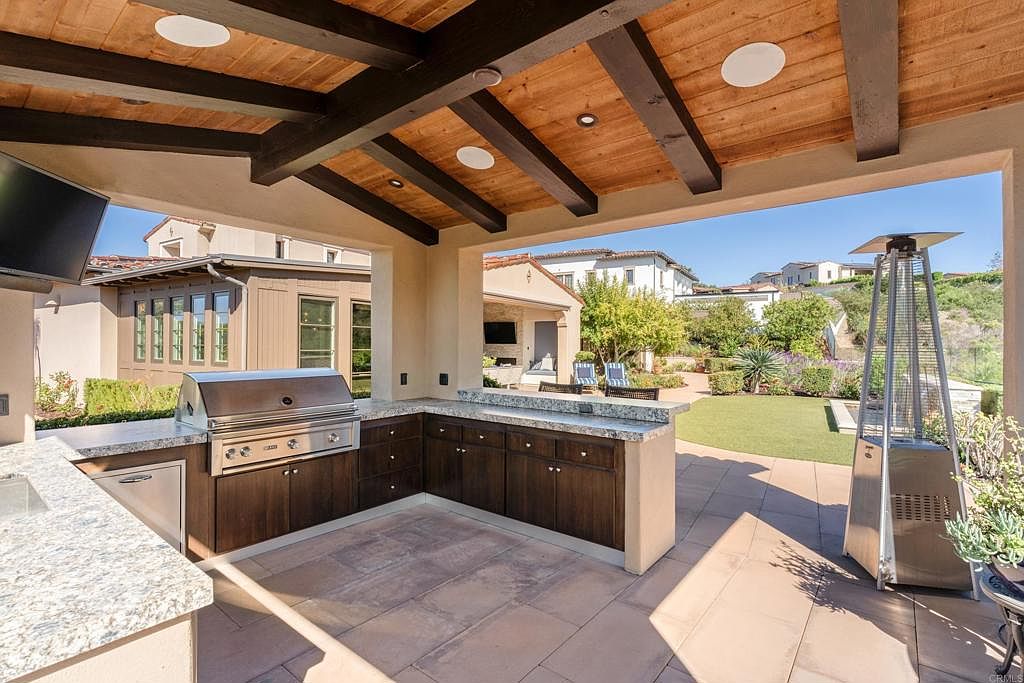 This image showcases an outdoor kitchen and patio area. The kitchen features dark wood cabinetry, granite countertops, and stainless steel appliances, including a built-in grill. The patio is paved with stone tiles and includes a tall, modern patio heater. The view extends to a well-maintained lawn and surrounding houses, creating an inviting outdoor living space.