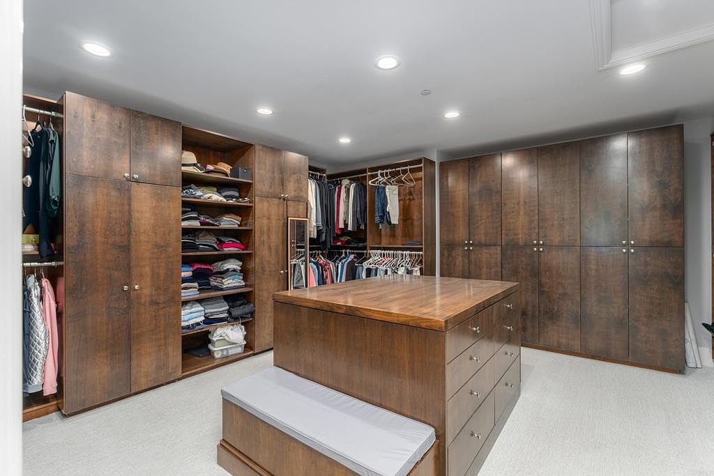 This is a spacious walk-in closet featuring custom wood cabinetry and shelving. An island with drawers and a cushioned bench sits in the center of the room, providing additional storage and seating. The closet is well-lit with recessed lighting, and the overall impression is one of organization and luxury.