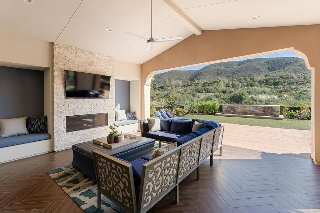 This image showcases a luxurious outdoor living space featuring a covered patio with a stylish seating area. The patio includes a comfortable blue sofa set with decorative back panels, a modern fireplace with a TV mounted above, and a view of a well-maintained lawn and distant hills. The flooring is a herringbone pattern, adding to the upscale feel of the space.